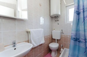 Et badeværelse med vask, toilet, badekar og et vindue med gardin.