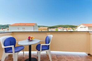 En balkon med to hvide stole og et rundt bord, en skål med frugt, og udsigt til huse og bakker i baggrunden.