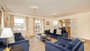 A living room with a blue sofa set, a glass coffee table, and a lamp. There are curtains on the windows, and a door leading to another room. - Riverside Home in Perth (Perth)