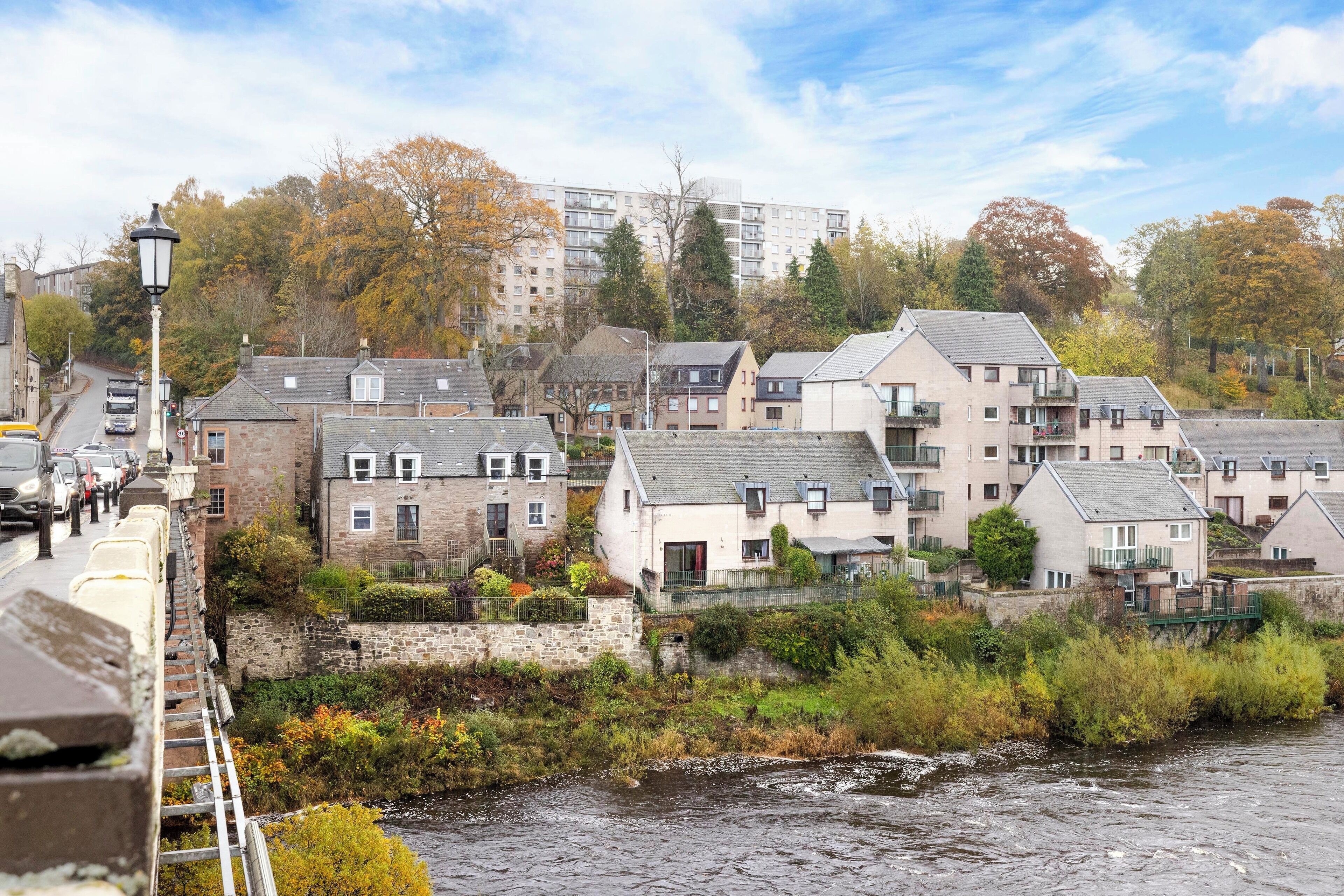 A riverside residential area with stone buildings, a bridge, and a modern apartment complex in the background.