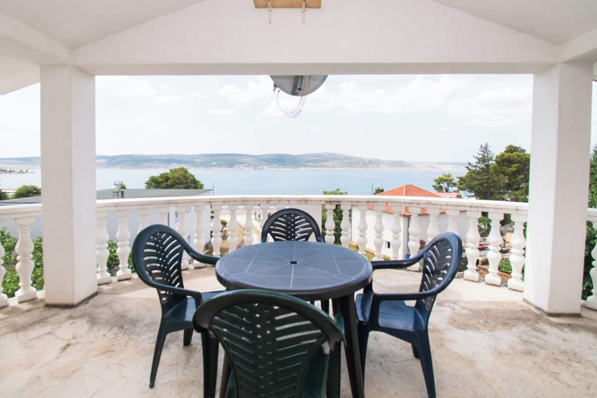 A covered outdoor seating area with a table and chairs, overlooking a body of water and distant land.