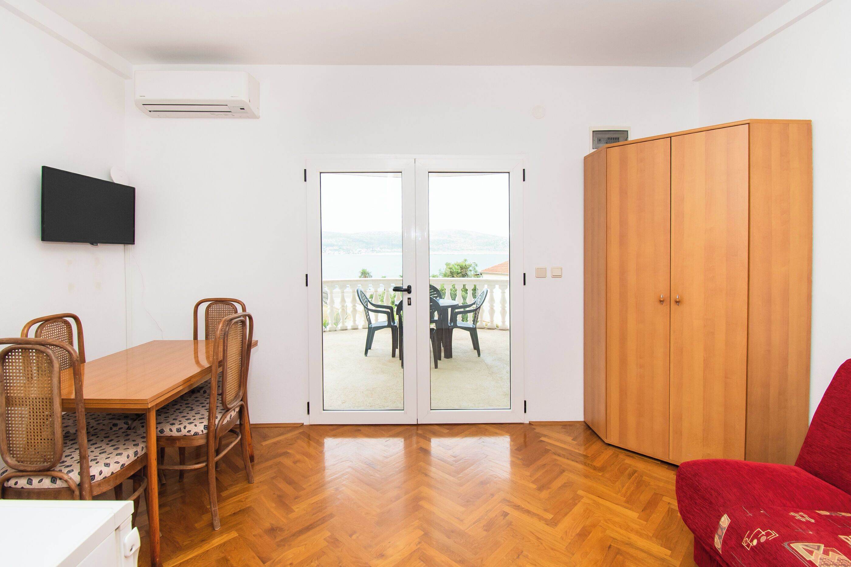 A room with a wooden herringbone floor, a dining table with chairs, a red sofa, a wooden wardrobe, and a sliding glass door leading to an outdoor area with a view of the sea.