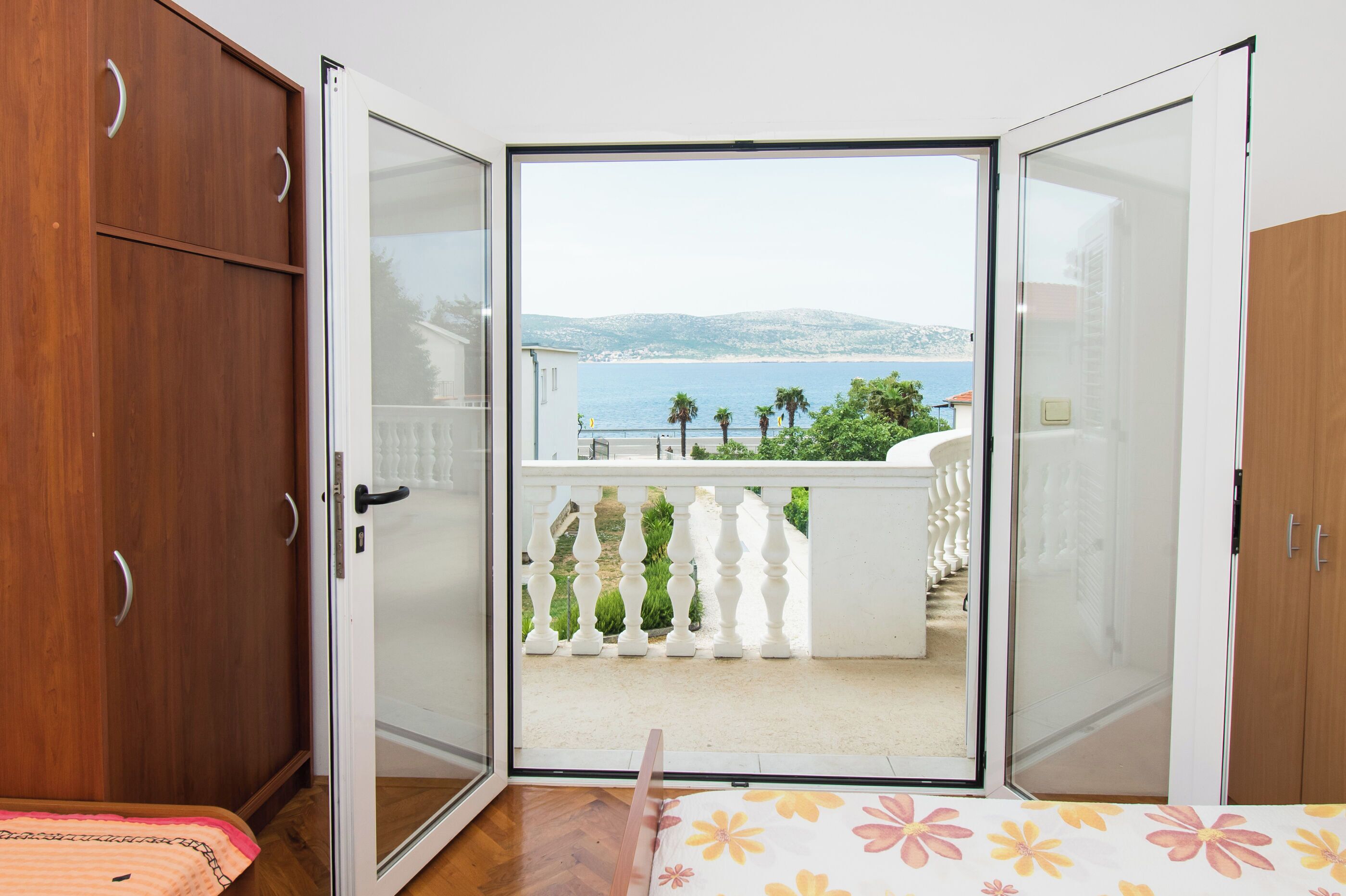 A room with a wooden wardrobe, a bed with a floral bedspread, and a balcony with a white balustrade and a view of the sea.