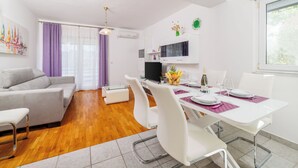 A modern living room with a sofa, dining table, and chairs. There is a TV, a small kitchen area, and a window with curtains. - Apartments Tanya (Rab)