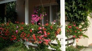 A covered porch with a white railing, surrounded by lush greenery and flowering plants. - Apartments Tanya (Rab)
