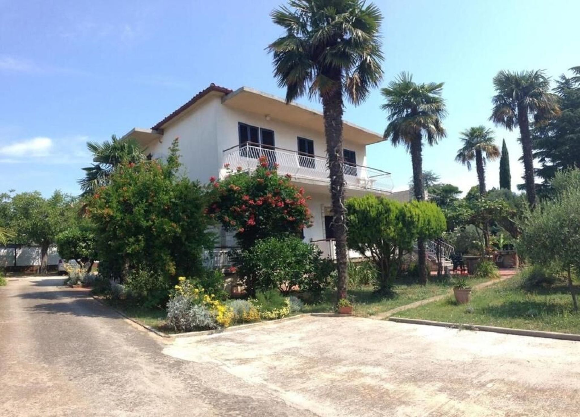 A two-story building with a balcony, surrounded by palm trees and various plants.
