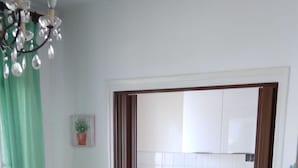 A room with a dining table, chairs, a bed, a chandelier, and a view of a kitchen through an open door. - Apartments Sea Breeze (Umag)