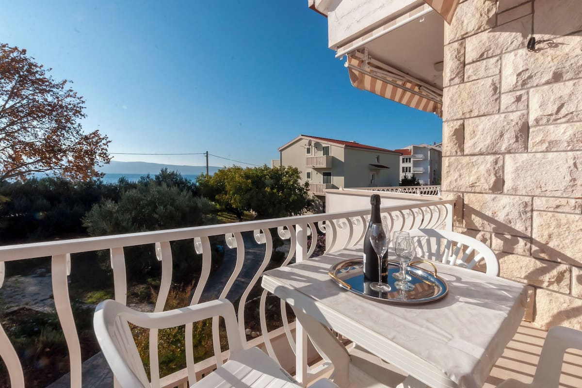 A balcony with a table set for two, overlooking a scenic view of a coastal area with buildings and trees.