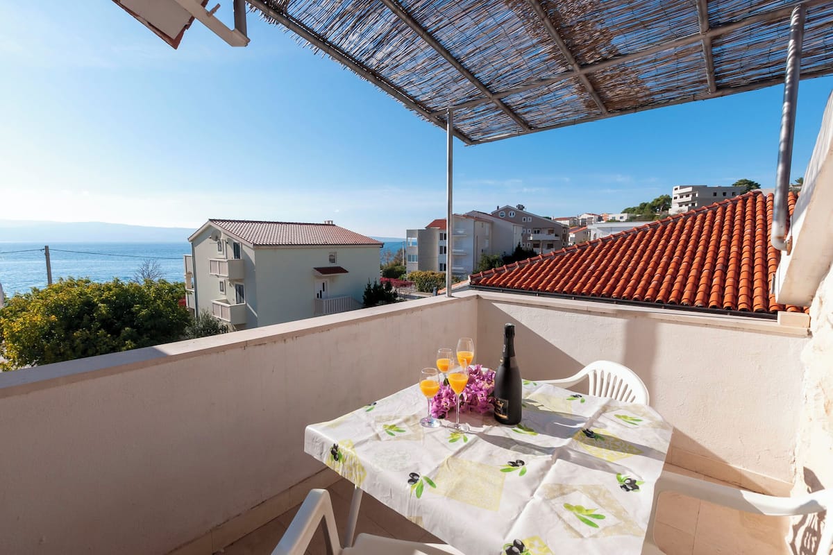 A balcony with a table set for two, overlooking a coastal view with buildings and a clear sky.