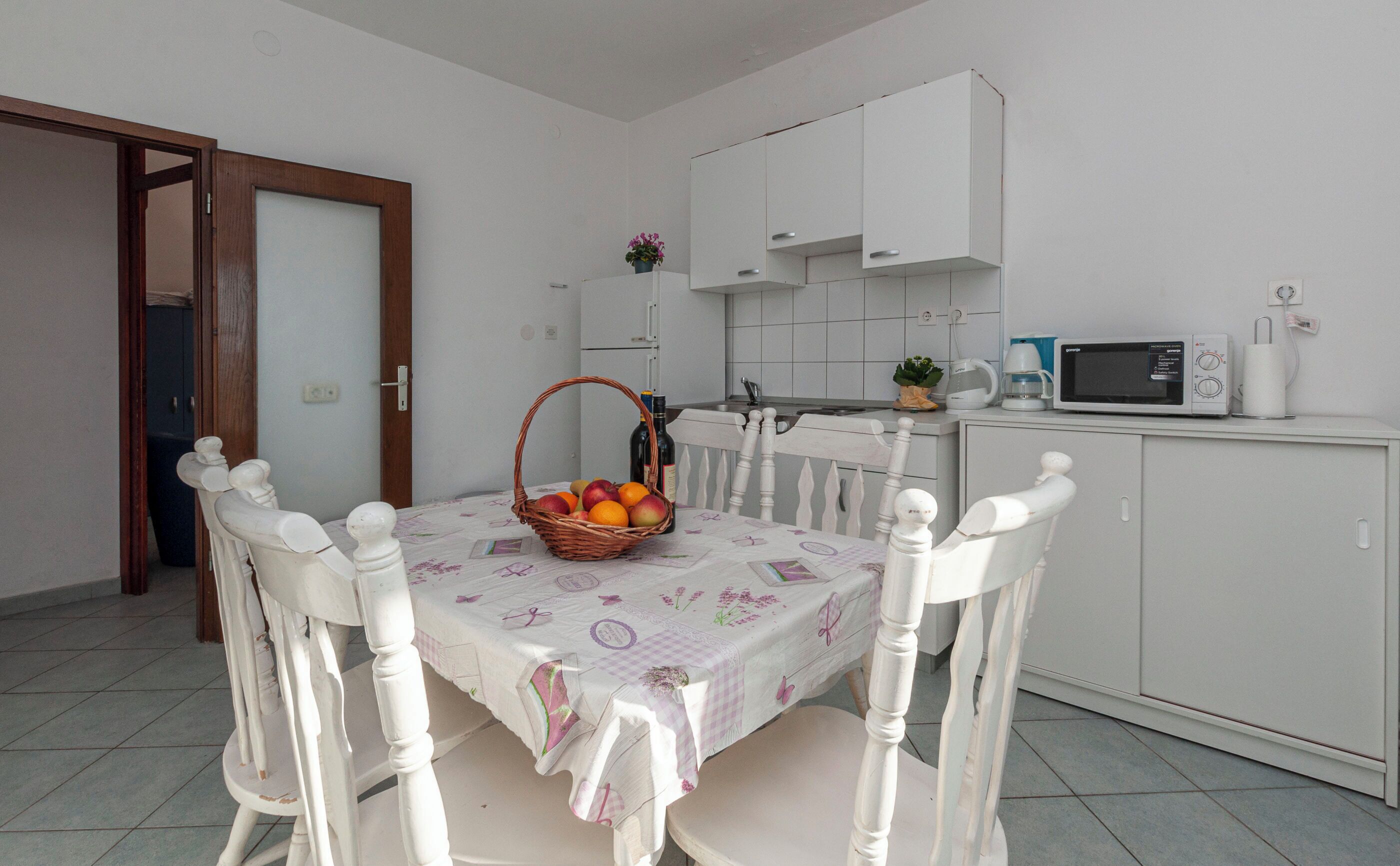 Un comedor con una mesa preparada para cuatro personas, rodeada de sillas blancas. Hay una cesta de fruta sobre la mesa y un mueble blanco con un microondas encima.