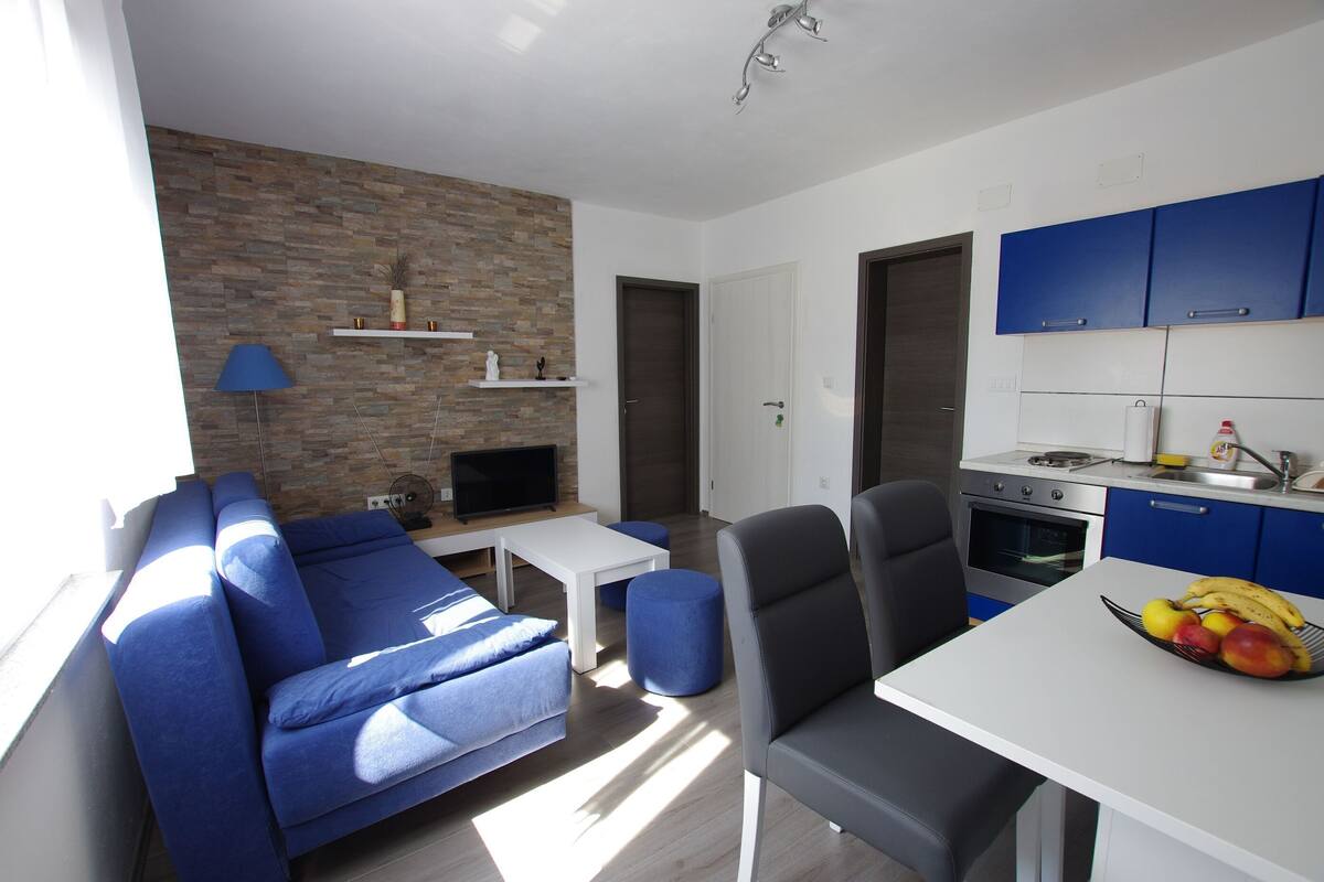Ein modernes Wohnzimmer mit einem blauen Sofa, einem kleinen Tisch und einem Fernseher. Es gibt eine Küchenzeile mit blauen Schränken und einer Obstschale auf der Arbeitsfläche.