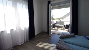 Una habitación con cama, puerta corrediza de vidrio a un espacio exterior con mesa y sillas, y cortinas translúcidas en las ventanas.