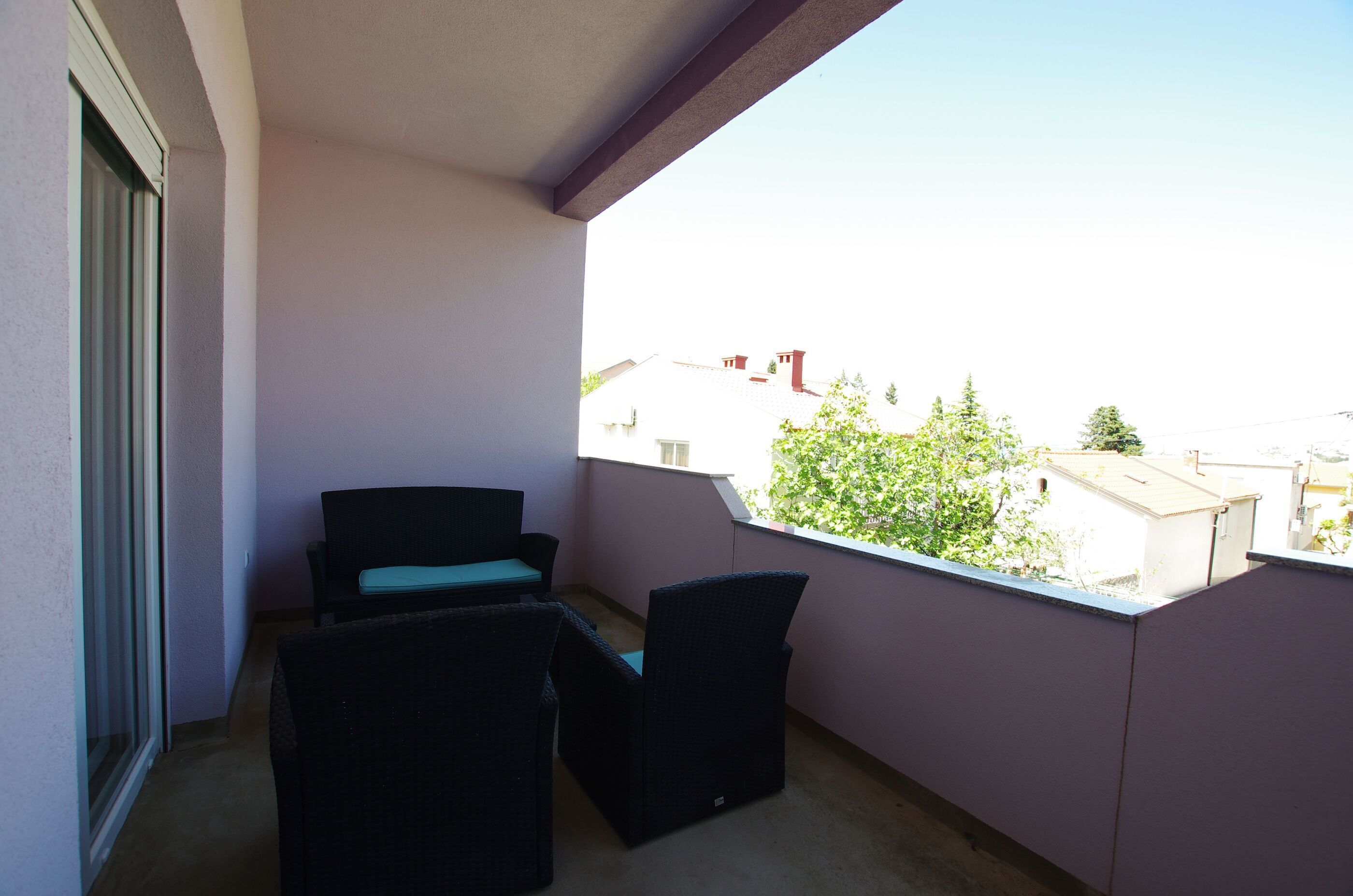 A balcony with two wicker chairs and a small table, overlooking a residential area with houses and trees.