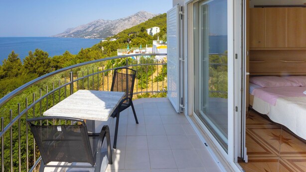 A balcony with a table and chairs, a bed, and a view of the sea and mountains. - Apartments Dream View (Brela)
