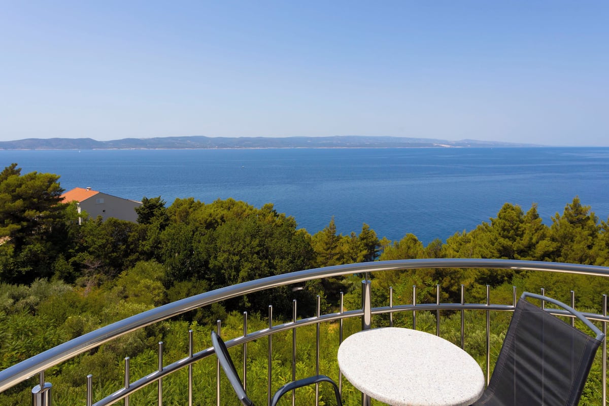 A balcony with a round table and chairs, overlooking a coastal landscape with trees and a view of the sea.