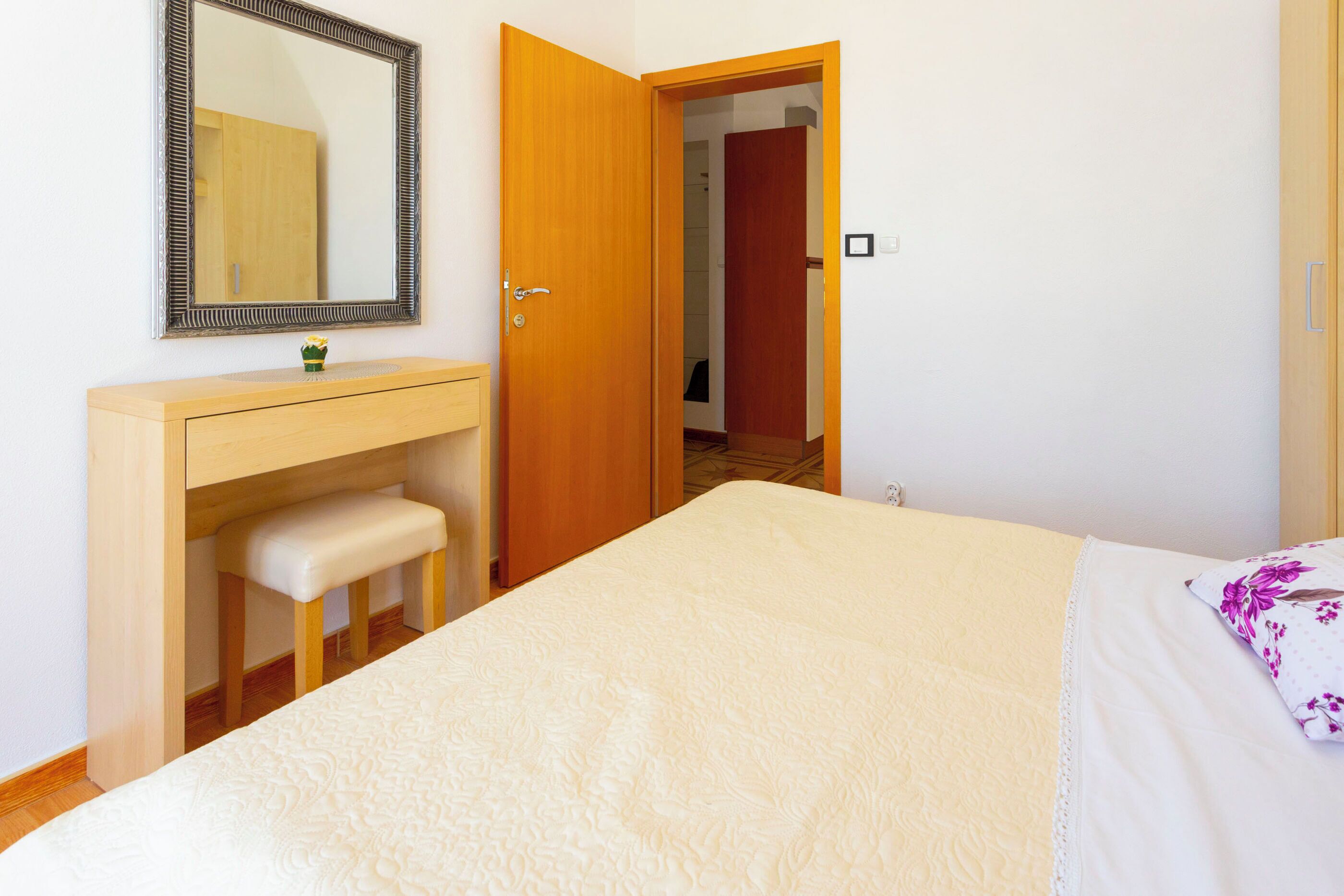 A bedroom with a bed, a wooden dresser, a bench, a mirror, and a door leading to another room.