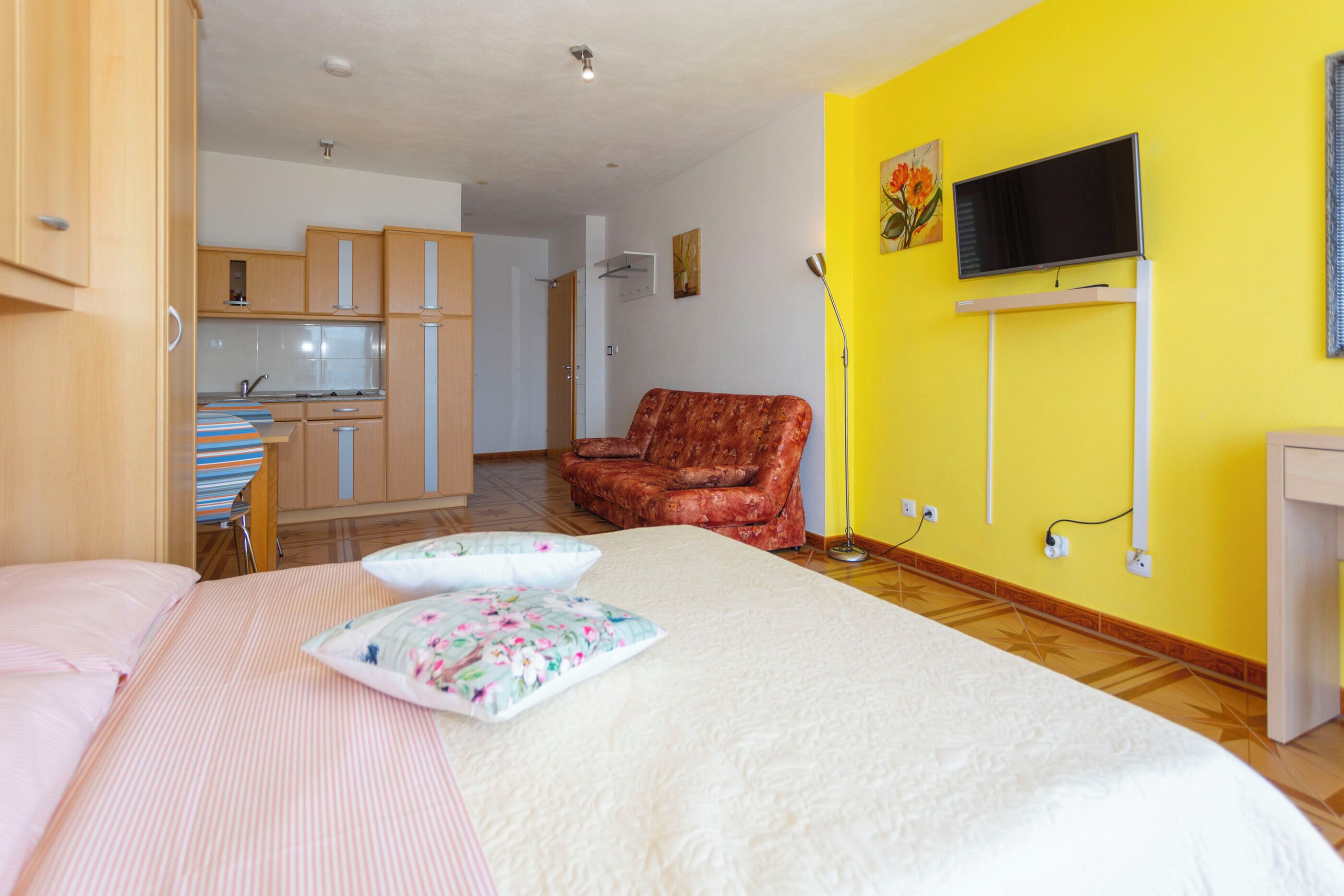 Una camera da letto con un letto, cuscini, una sedia, una TV e una cucina sullo sfondo.