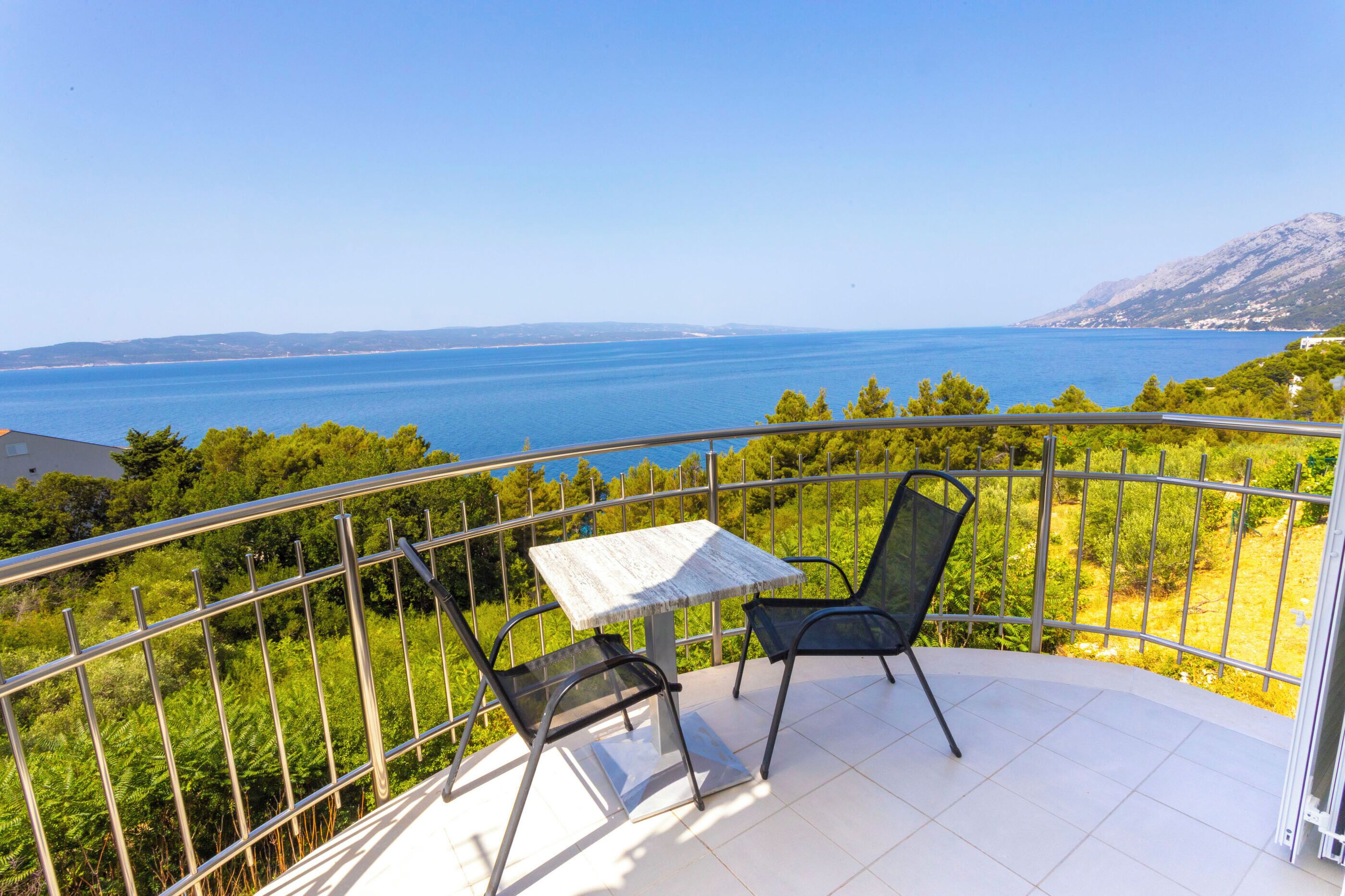Un balcon avec une table et deux chaises, offrant une vue dégagée sur un paysage côtier, avec la mer et des collines lointaines en toile de fond.