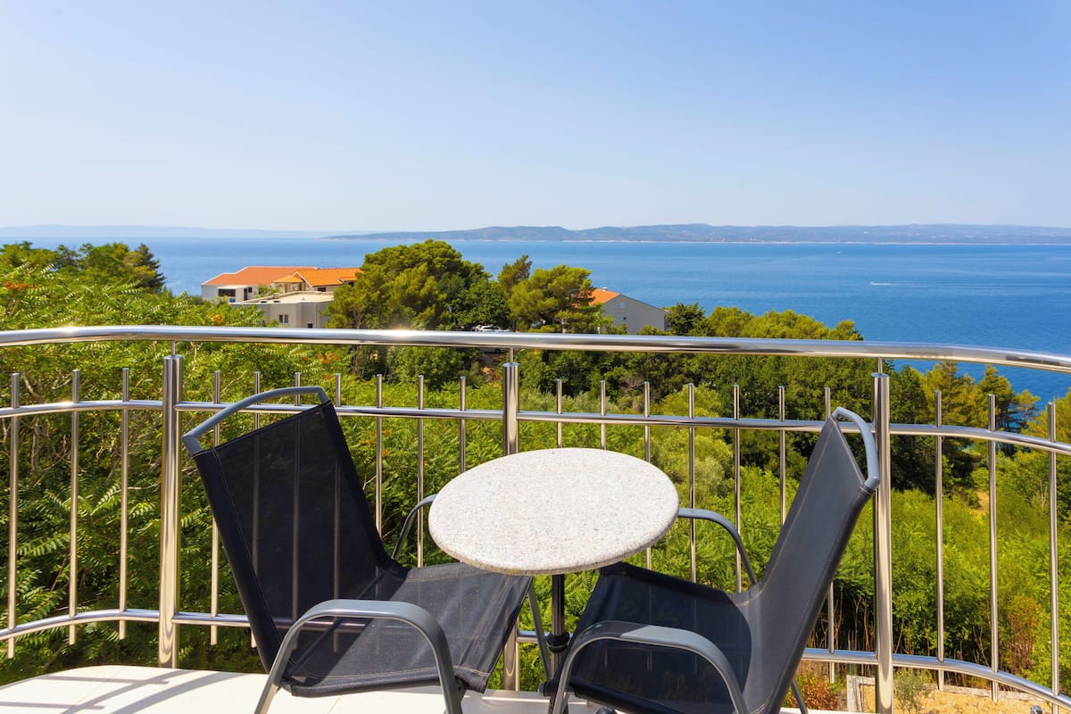 A balcony with two chairs and a small table, overlooking a coastal view with houses and trees.