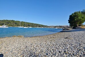 Ein Kiesstrand mit Booten im Wasser und einem Baum rechts.
