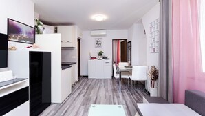 Μια σύγχρονη κουζίνα με λευκά ντουλάπια, μια τραπεζαρία με ένα λευκό τραπέζι και καρέκλες, και ένα καθιστικό με έναν καναπέ και μια τηλεόραση.