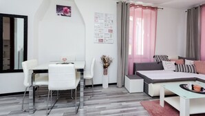 Ένας μοντέρνος, καλά φωτισμέν ος χώρος με τραπεζαρία, ένα κρεβάτι με γκρι και λευκούς τόνους και ένα μεγάλο παράθυρο με κουρτίνες.