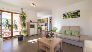 Een moderne woonkamer met een bank, een bijzettafeltje met een bloevas, en uitzicht op de keuken en het balkon.