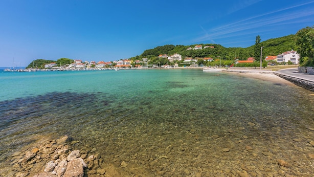 A coastal town with clear waters, rocky beach, and buildings on a hillside. - Apartments Marija G (Rab)