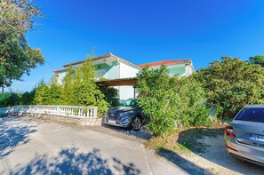 Uma casa de dois andares com telhado de telhas vermelhas, uma entrada para carros e dois veículos estacionados.