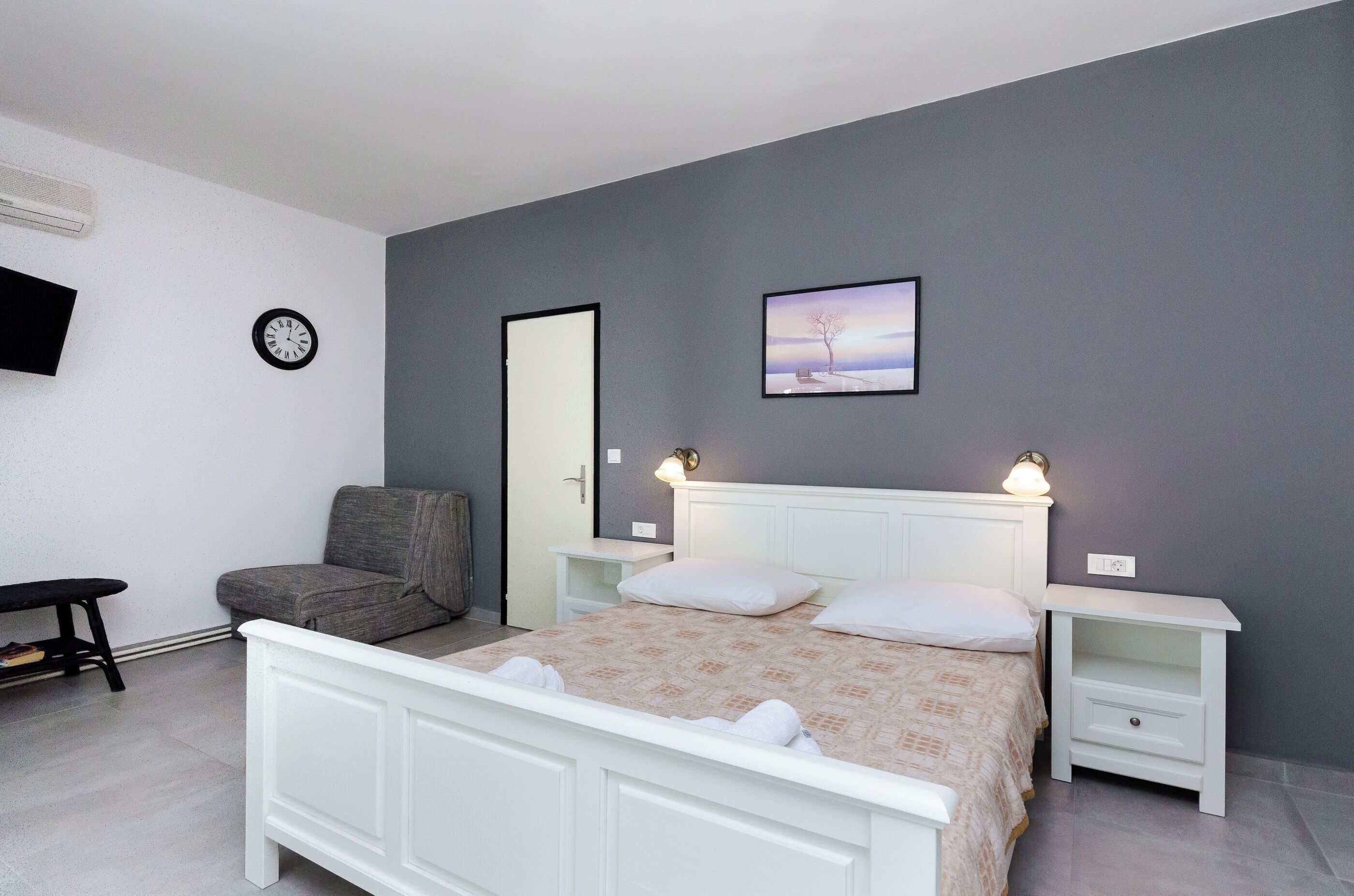 Un dormitorio con cama, mesitas de noche, una silla, un reloj y un cuadro en la pared.