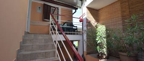 Una escalera con barandillas rojas, una planta en maceta y una ventana con contraventanas marrones.