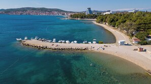 Eine Luftaufnahme einer Küstenlandschaft mit Sandstrand, Booten und einer Stadt am Horizont.