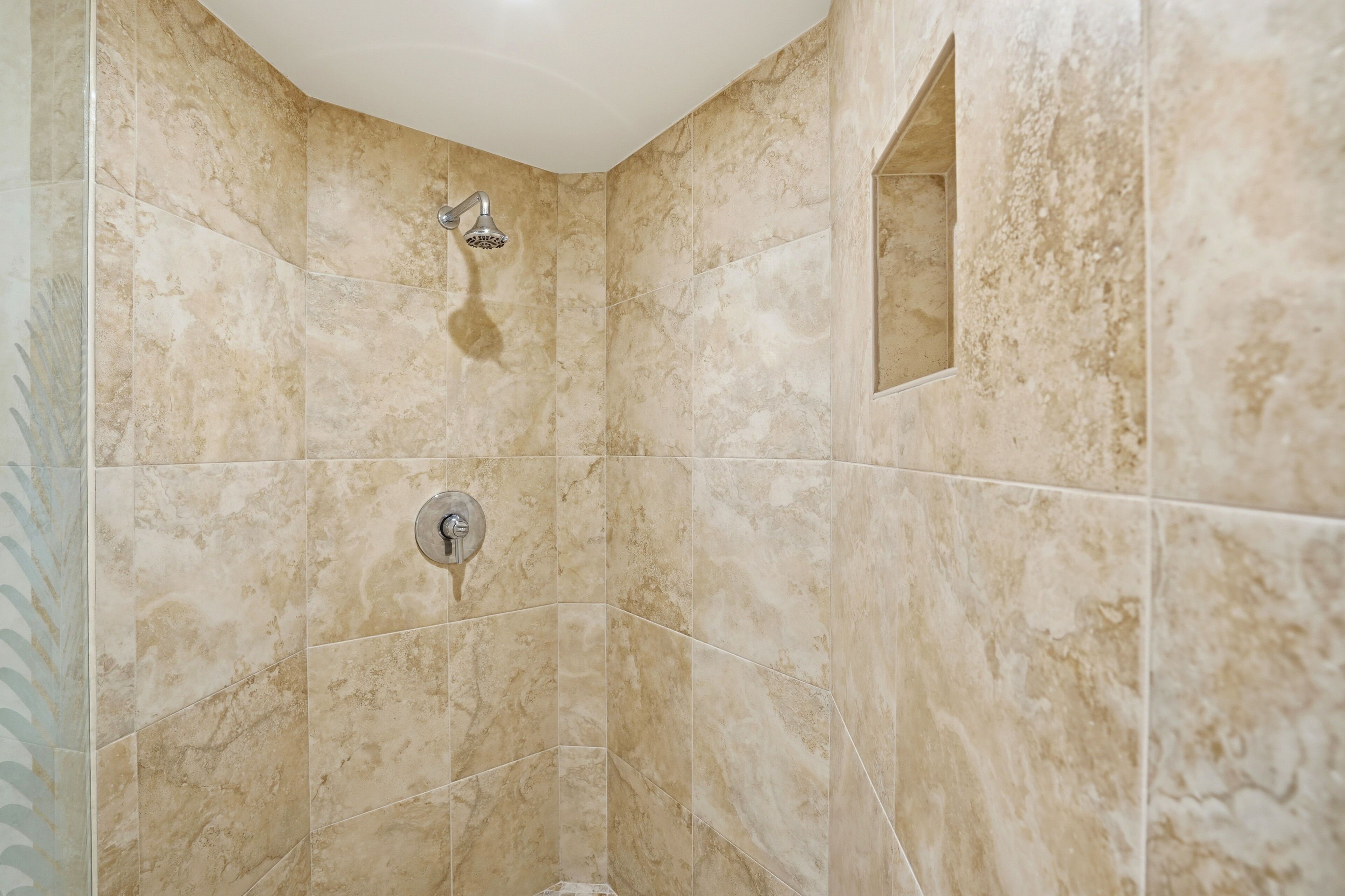 Une douche d’angle avec des carreaux de marbre beige, un pommeau de douche et un petit miroir.