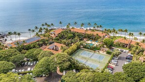 Ett flygbildsfoto av ett semesterområde med en strand, palmer, solpaneler på byggnaderna, en tennisbana och en parkeringsplats.