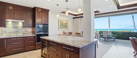 A modern kitchen with dark wood cabinets, a marble countertop, and a large island. There is a dining area with a view of the ocean.