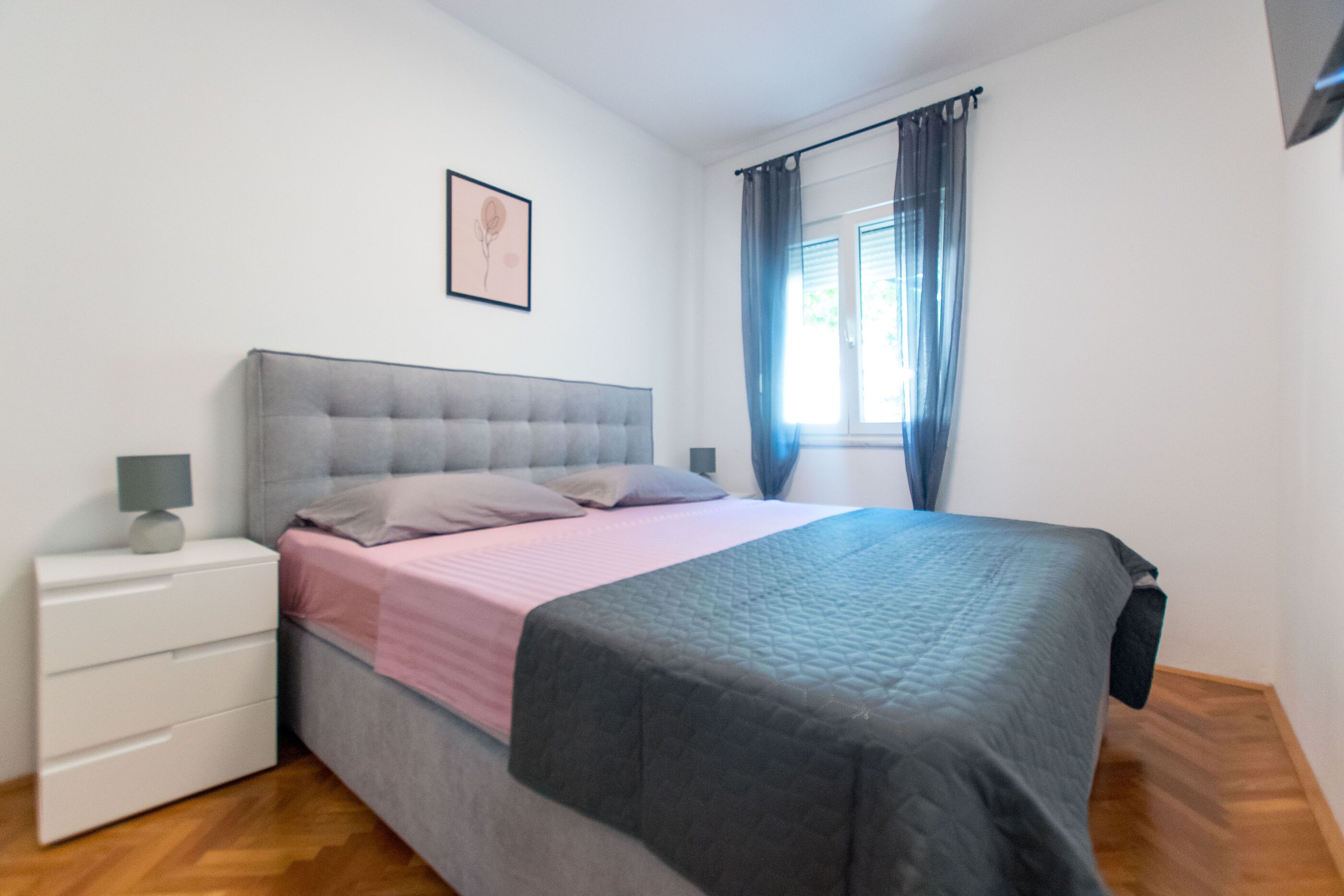 A bedroom with a bed, a nightstand, a lamp, a window with curtains, and a framed picture on the wall.