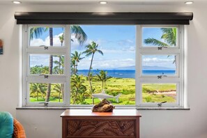 A room with a large window offering a view of palm trees and a beach. - Renovated 2BR w/ AC - Menehune Shores #522 Oceanview South Maui Condo Getaway (Kihei, Maui)