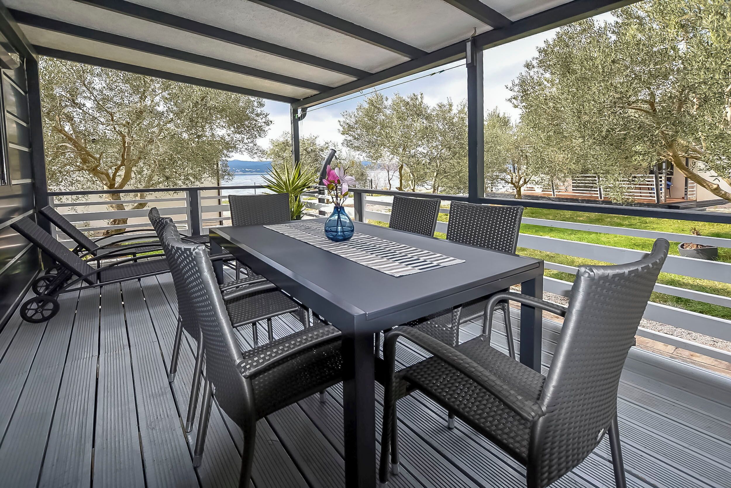 Une terrasse extérieure avec une table et des chaises, un vase rempli de fleurs et une vue sur des arbres et une étendue d’eau.