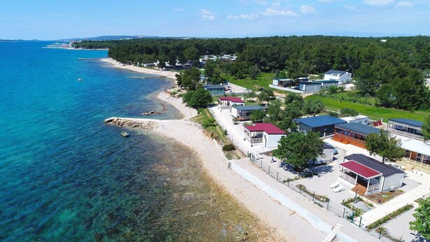 An aerial view of a coastal area with a beach, several buildings, and a forested hill in the background. - Mobile Home Pen Shell (Biograd na Moru)