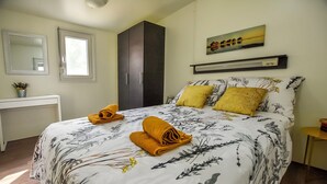 A neatly made bed with floral bedding and two yellow pillows, a dark wardrobe, a small bedside table with a lamp, a window with a view, and a painting on the wall. - Mobile Home Pen Shell (Biograd na Moru)