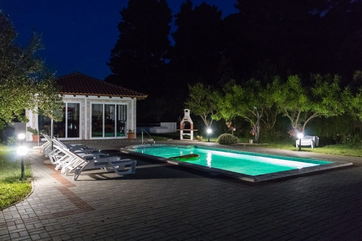 Ein Schwimmbecken mit Liegestühlen und Grillplatz, umgeben von Bäumen und einem Haus mit einem Ziegeldach.