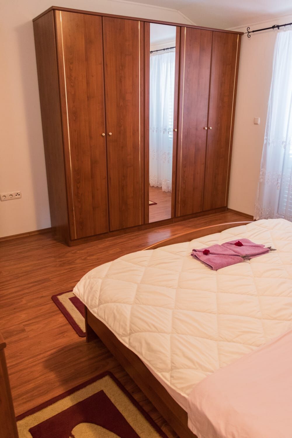 Een slaapkamer met een grote houten garderobe, een bed met quilt en een spiegel die de kamer weerspiegelt.