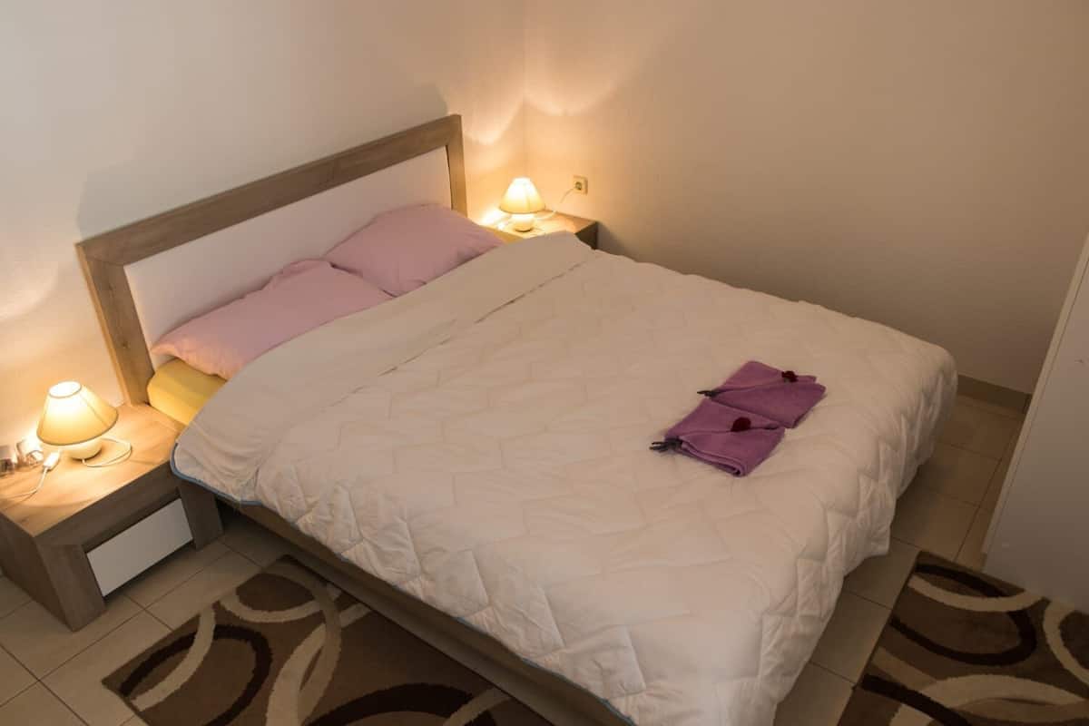 A neatly made bed with a white quilt, two pink pillows, and a purple towel on the side. There are two bedside lamps with white shades.