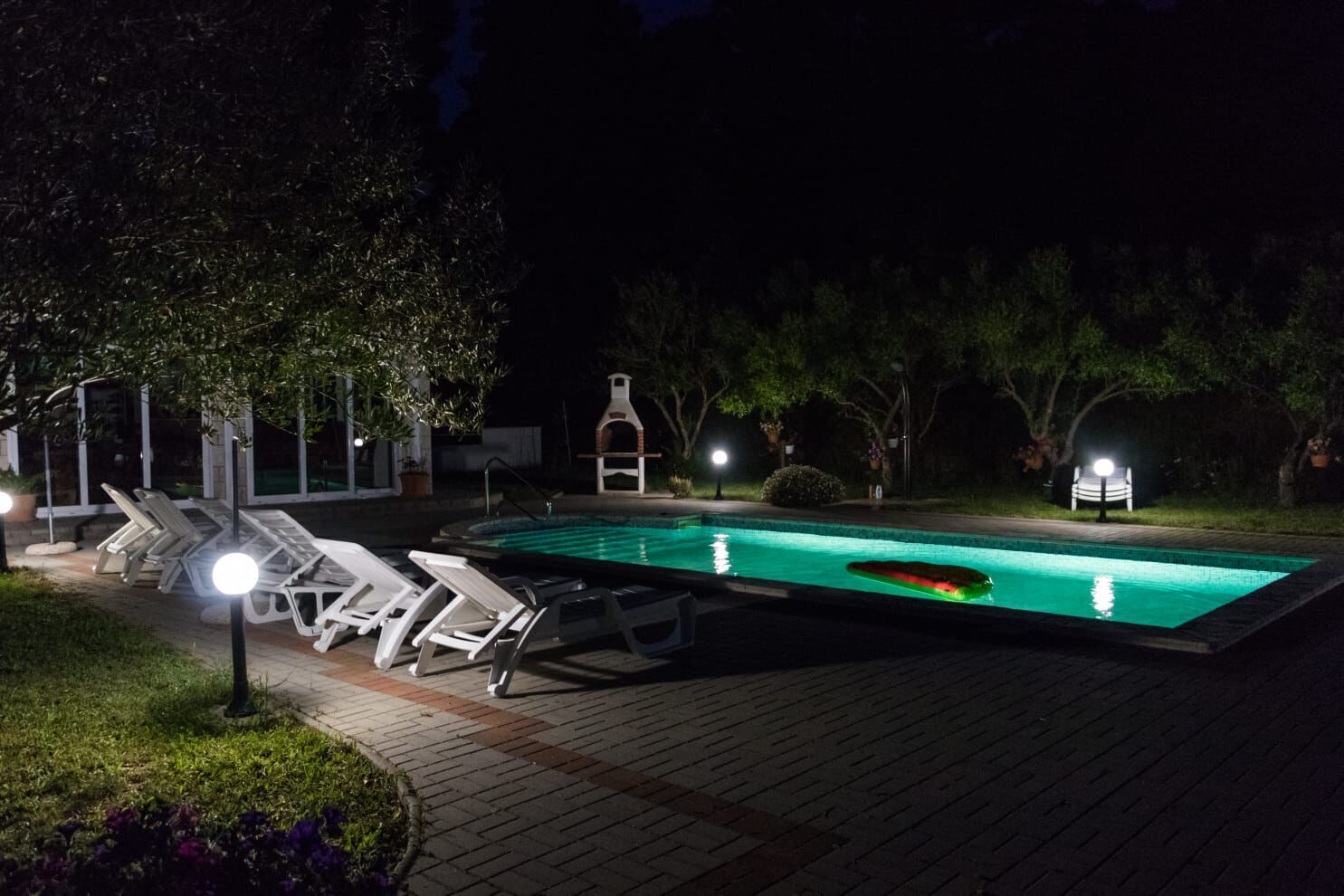 A swimming pool with lounge chairs and a barbecue area, surrounded by trees and illuminated by night lights.