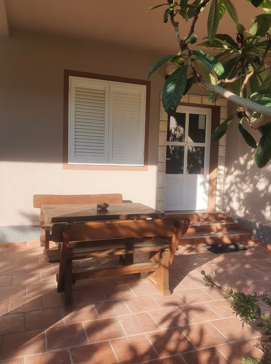 A patio with a wooden table and benches, a door, and a window with shutters.