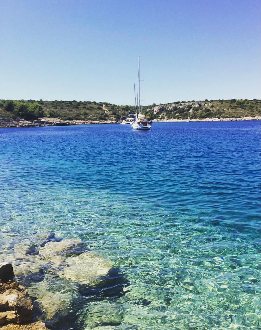 A clear blue sea with a sailboat in the distance and rocky shorelines.