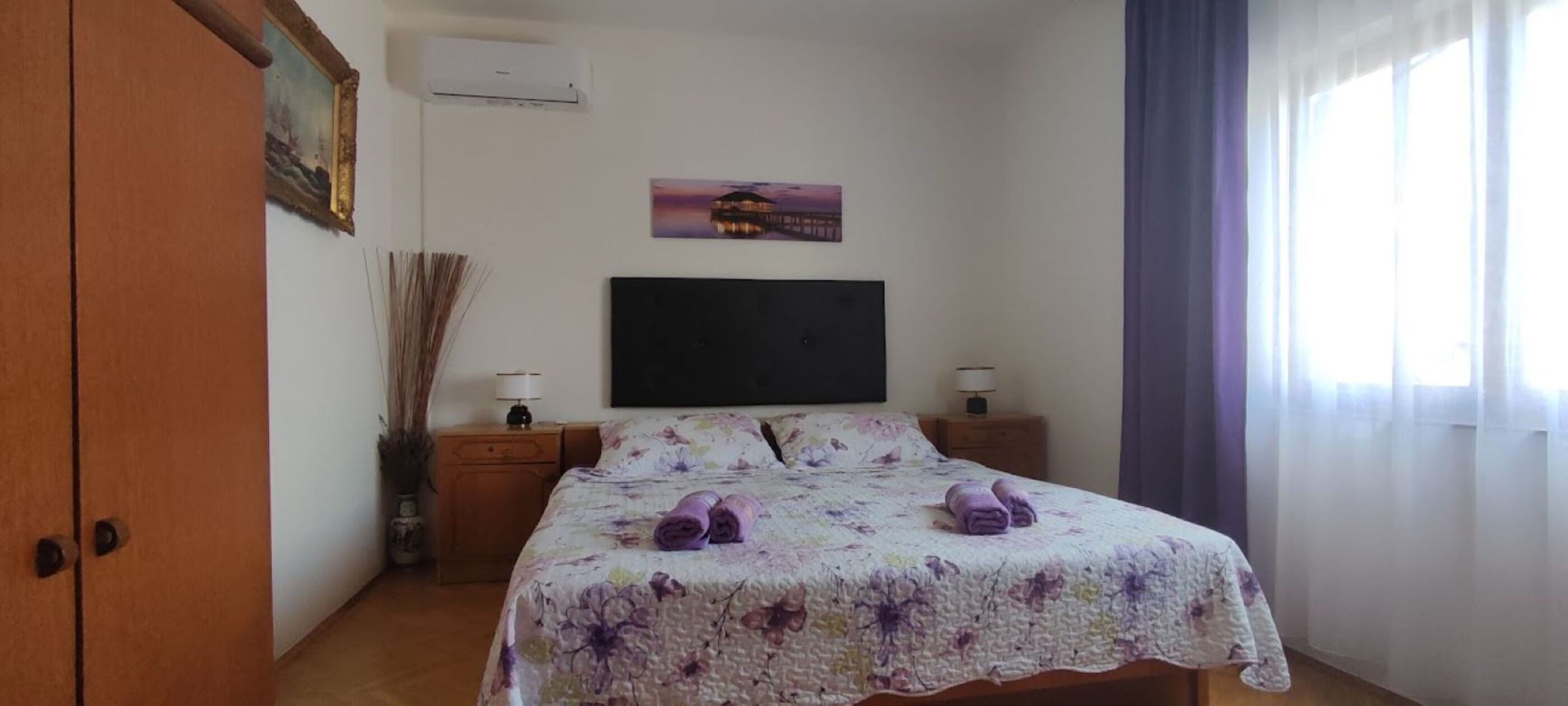 Une chambre à coucher avec un lit, deux tables de chevet, un téléviseur fixé au mur et une fenêtre avec des rideaux.