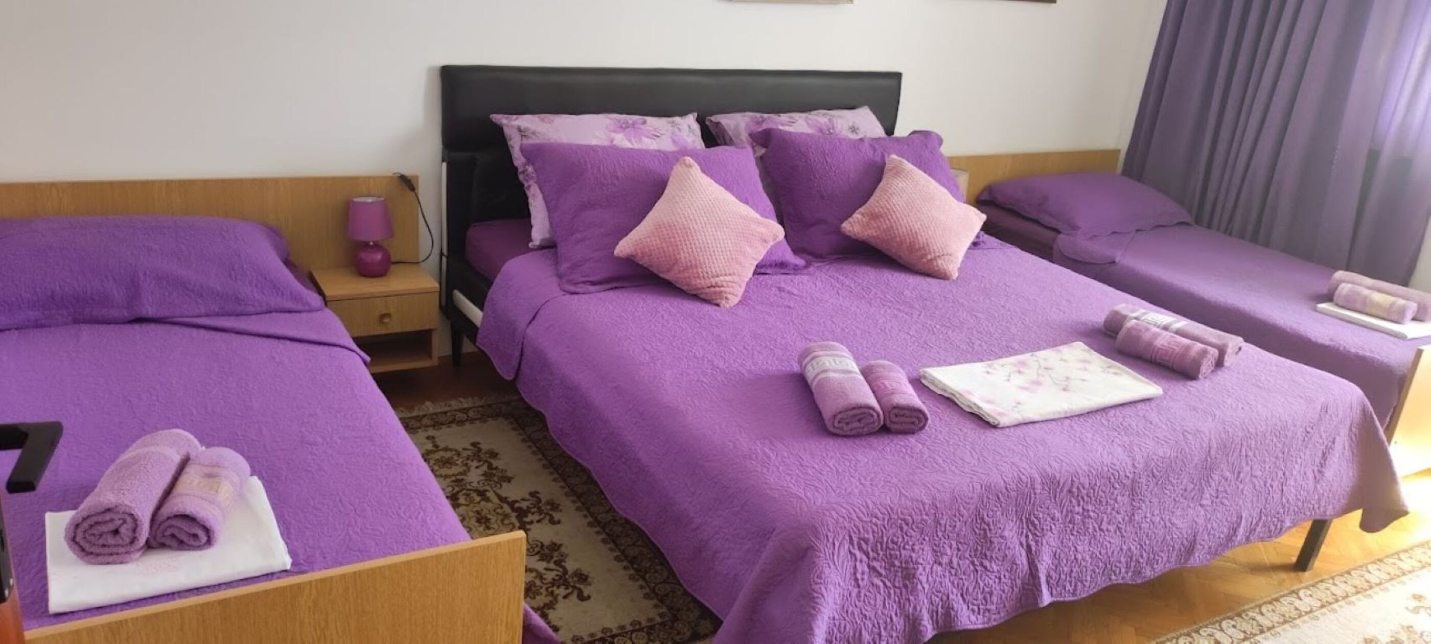 A hotel room with two beds, each with purple bedding and pillows. There are towels on the beds and a small table with a lamp.
