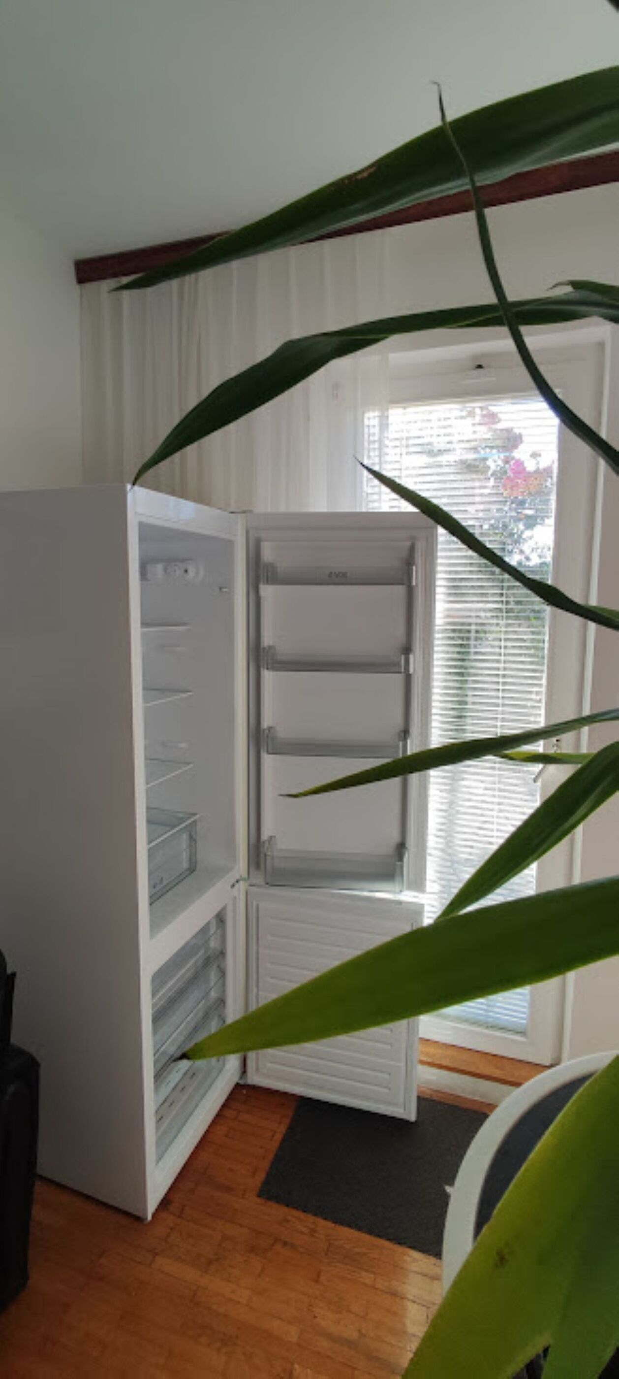 An open refrigerator with multiple shelves and a freezer compartment, positioned in a room with a wooden floor and a window with blinds.