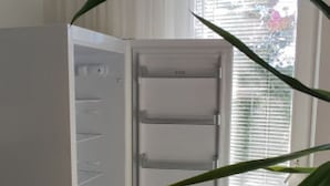 Un refrigerador abierto con varios estantes y un compartimento congelador, ubicado en una habitación con piso de madera y una ventana con persianas.